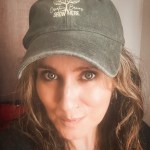 A close-up portrait of a woman wearing a dark cap with the text 'Creative Brains GROW HERE' and a brain graphic, featuring wavy hair and a natural makeup look.